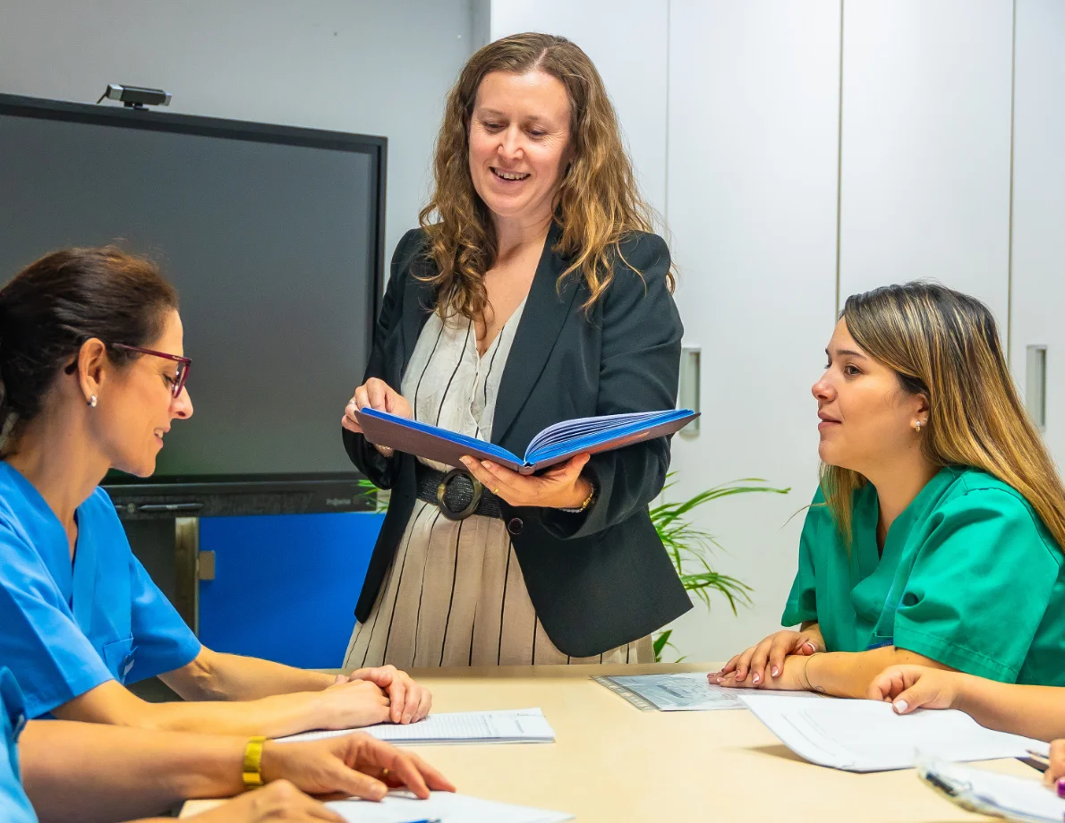 A woman stands holding an open binder, conducting staff training for two women in scrubs seated at a care home table.
